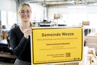 Irina Schultz von der Wirtschaftsförderung Kreis Kleve präsentiert das Ortsschild der Gemeinde Weeze mit der Einladung zum Unternehmerfrühstück. 