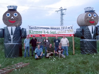 Mitglieder des Tambourcorps Weeze vor den Strohpuppen, die auf die diesjährige Kirmes jeweils an den Ortseingängen hinweisen