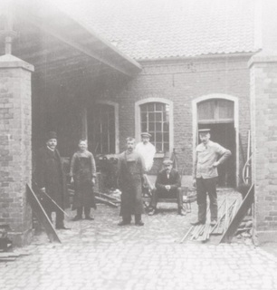 'Alte Schmiede' (old blacksmith’s). The two bricked gate posts are still visible today. The left half of the yard was covered by a roof, this is where the shodding stall stood and is where the horses were shod. The workplace was expanded in the 1920’s, as is still visible today. From left to right: Heinrich Belting (Weeze’s coppersmith and plumber), two unknown journeymen (trainees), Theodor Weigand (master painter), Heinrich Verhülsdonk (owner of the pub on Kirchplatz/corner Kirchplatz/corner Loëstrasse) and blacksmith Theodor Kerns, postcard-view from around 1910.